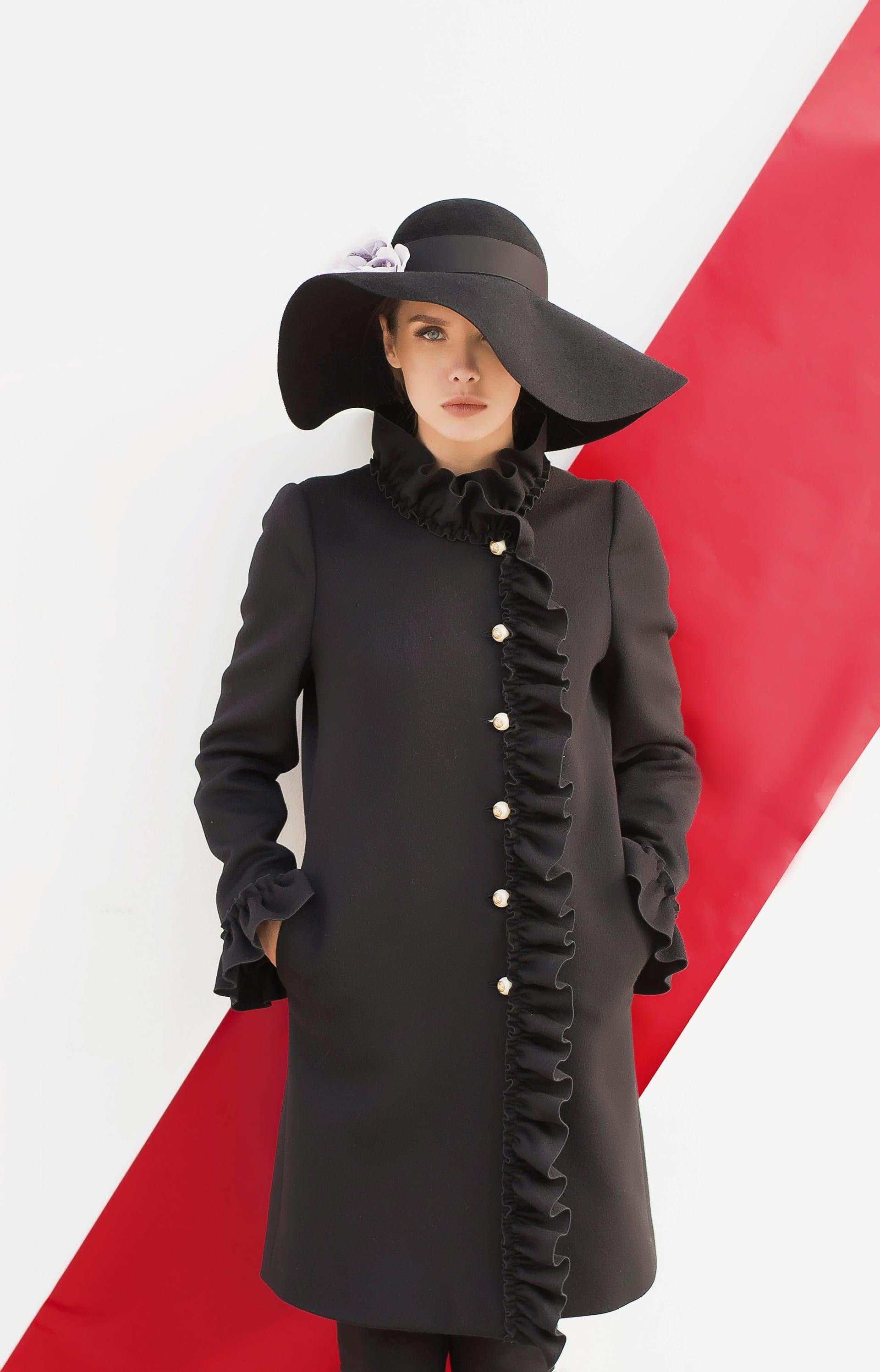Model in a black ruffled coat and wide-brimmed hat against a red and white backdrop